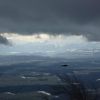 Bild um 11:48 mit einer vorbeifliegenden Dohle, dem Mittelland und den leuchtenden Alpen in Föhnlage. Die schwarzen Wolken werden vom Sturm rasend schnell vorbei getrieben.